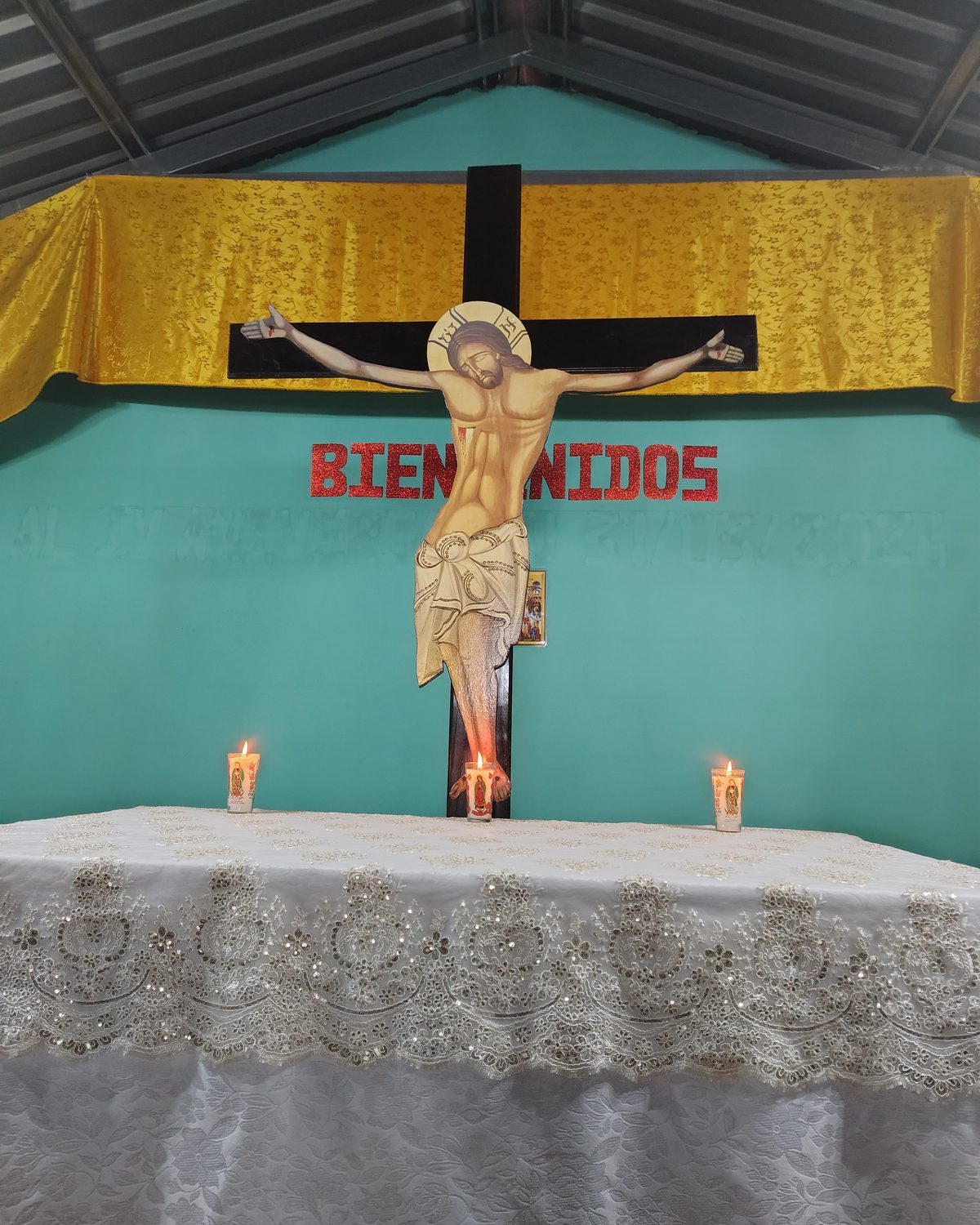 A parish church today: a crucifix and the words Bien venidos on a turquoise wall, above a single table with two small candles